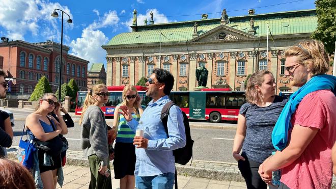 &ldquo;a man wearing sunglasses surrounded by a group of tourists across from a neoclassical building&rdquo;